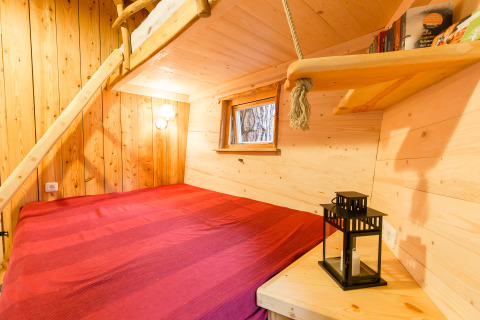 Cabane dans les arbres chaleureuse avec lit rouge, murs en bois, livres et lanterne près de la fenêtre.