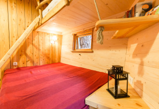 Indbydende træloungelignende boomhut med rødt sengetæppe, trævægge og hyggelig vinduesudsigt.