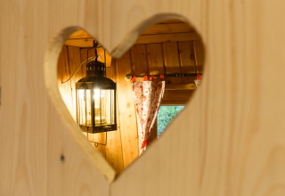 Uitzicht door een hartvormige uitsparing in houten wand op lantaarn en gordijnen in TreeLounge - Boomhut Nedersaksen.