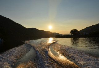 Solnedgang over Mosel-floden set fra en båd, med vandets spor oplyst og grønne bakker i baggrunden.