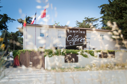 Ingresso al Camping Les Cyprès - Glampingtenten Vendée con bandiere, edificio e alberi sullo sfondo.