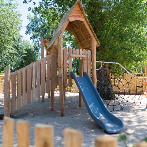 Parque infantil de madera con tobogán y red de escalar en Camping Les Cyprès - Glampingtenten Vendée bajo los árboles.
