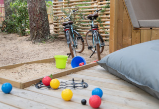 Udsigt over glampingtelt terrasse med legeplads, sandkasse, legetøj og cykler på Camping Les Cyprès.