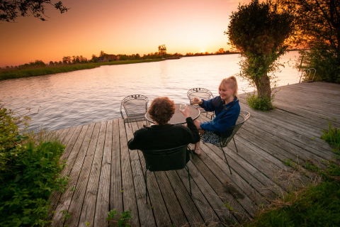 Zwei Personen entspannen mit Getränken auf einer Holzterrasse am Wasser bei Sonnenuntergang bei Pean Buiten Friesland.