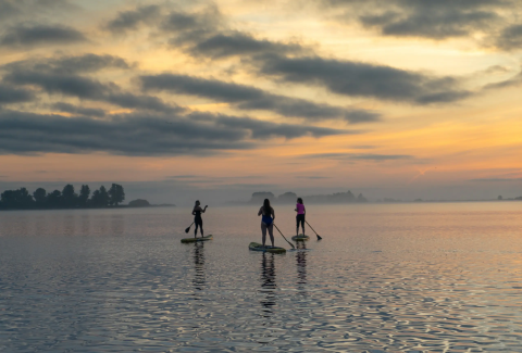 Drie mensen suppen op een rustige plas bij zonsondergang nabij Pean Buiten - Natuurhuisjes Friesland.