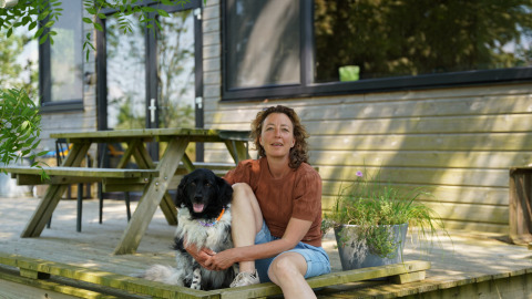 Femme assise avec son chien sur une terrasse en bois devant un chalet à Pean Buiten - Natuurhuisjes Friesland.