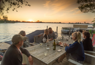 Eine Gruppe sitzt am Holztisch am Wasser bei Sonnenuntergang im Pean Buiten - Natuurhuisjes Friesland.