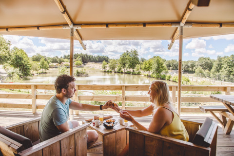 Couple enjoying breakfast on a terrace overlooking a lake at Camping Village de la Guyonnière glamping site.