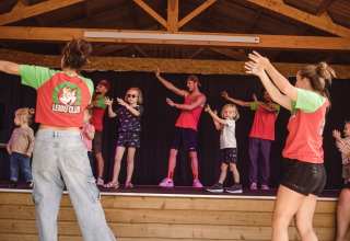 Børn og voksne deltager i en danseaktivitet på scenen i Lenny Club ved Camping Village de la Guyonnière.