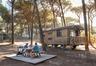 Family sits at picnic table by wooden glamping cabin at Huttopia Parque de Doñana – Glamping Andalusia.