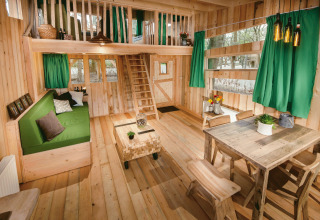 Interior view of a treehouse with green curtains, rustic wooden furniture, and a loft bedroom at Boomhut Brabant.