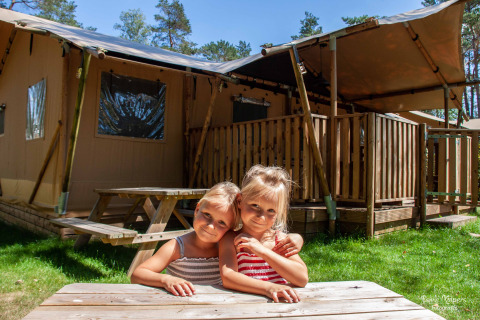 To børn hygger sig ved et træbord foran glamping-safarilodge i skoven i Gelderland på en solskinsdag.