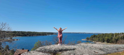 Person stands with arms raised on a rock overlooking the water at Sävö Bed and Breakfast Glampingtenten Södermanland.