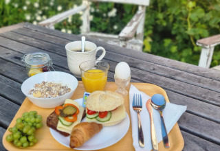 Desayuno al aire libre sobre mesa de madera con pan, croissant, fruta, jugo y café durante el glamping.