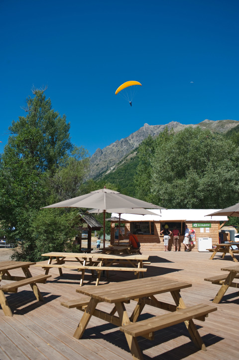 Udendørs opholdsområde med træborde, parasoller og udsigt til paraglider nær Huttopia Vallouise, Hautes-Alpes.