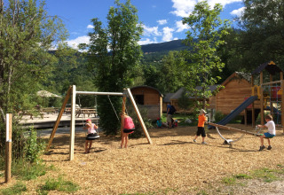Niños juegan en el área de juegos de Huttopia Vallouise - Glamping Hautes-Alpes, rodeados de cabañas y naturaleza.