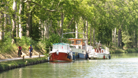 Twee fietsers rijden langs boten aan een rustige kanaal bij Nature's Pace Holidays - Glamping Occitanie.