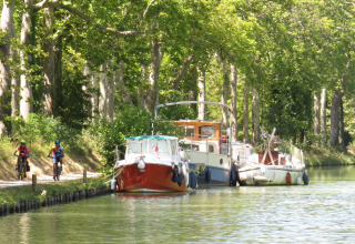 Twee fietsers rijden langs boten aan een rustige kanaal bij Nature's Pace Holidays - Glamping Occitanie.