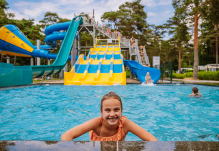 Niña en la piscina sonriente con toboganes coloridos en Camping Samoza – Glamping Lodges Veluwe rodeado de bosque.