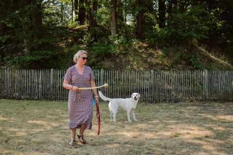 Frau spielt mit Hund auf einer Wiese vor Zaun und Bäumen bei Camping Samoza – Glamping Lodges Veluwe.