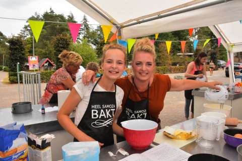 Deux femmes cuisinent lors d'un concours de pâtisserie extérieur à Camping Samoza – Glamping Lodges Veluwe, décoré de fanions.