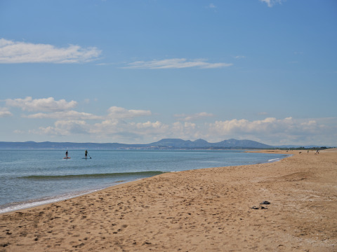 Peaceful sandy beach at Camping Nautic Almata – Glamping Costa Brava with paddleboarders and mountains in view.