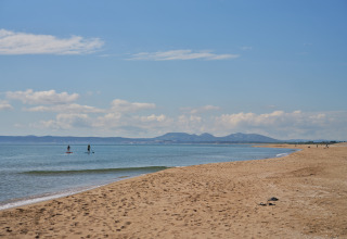 Rolig strandudsigt ved Camping Nautic Almata – Glamping Costa Brava med paddleboardere og bjerge i baggrunden.