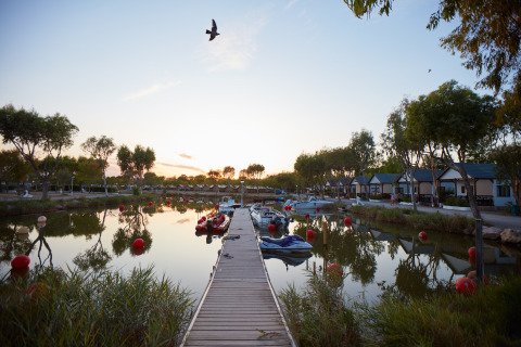 Camping Nautic Almata – Glamping Costa Brava avec ponton, bateaux, cabanes et arbres au bord de l’eau au coucher du soleil.