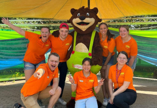 Gruppe af unge medarbejdere i orange skjorter poserer med maskot på Camping Beringerzand - Finse Kota's Limburg.
