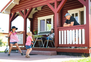 Gezin geniet bij een houten huisje in Ostseecamping Zierow terwijl kinderen vrolijk op het terras spelen.