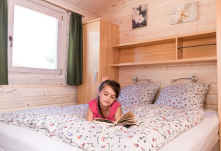 Niña leyendo un libro en una cama con sábanas florales en una cabaña del Ostseecamping Zierow, Alemania.