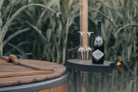 Champagne bottle and two glasses beside a wooden tub surrounded by greenery at Glamping La Ferme.