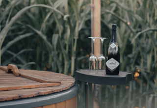 Bouteille de champagne et deux verres près d’une cuve en bois entourée de verdure au Glamping La Ferme.