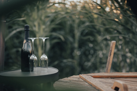 Eine Champagnerflasche und zwei umgedrehte Gläser stehen auf einem Tisch neben einem Holzstuhl im Grünen.
