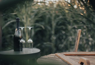 Een fles champagne en twee omgekeerde glazen op een tafel naast een houten stoel in een groene buitenomgeving.