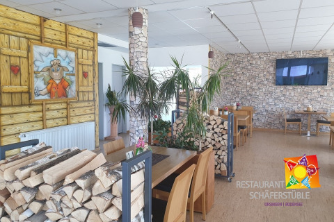 Interior de restaurante con leña apilada, mesas de madera, paredes de piedra y una pintura de una oveja.