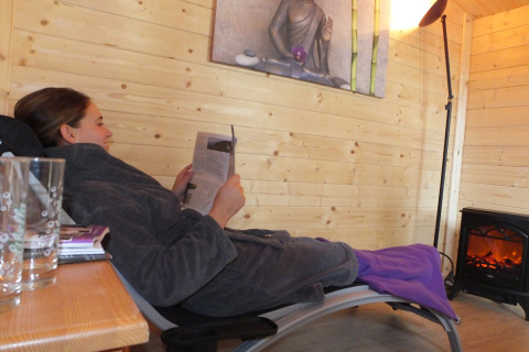 Mujer relajándose en una tumbona leyendo una revista en una acogedora cabaña de madera en el camping Orsingen.