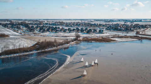 Lago ghiacciato con barche a vela davanti a lodge di lusso e paesaggio innevato a Friese Meren Villa’s, Frisia.