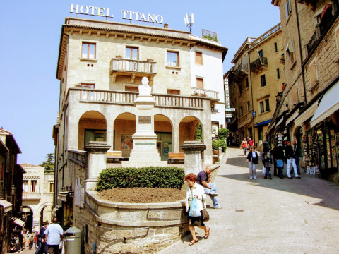 Historisch hotel in het centrum van San Marino, mensen lopen op een zonnige dag langs winkels en restaurants.