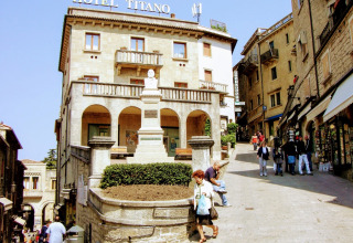 Historisch hotel in het centrum van San Marino, mensen lopen op een zonnige dag langs winkels en restaurants.