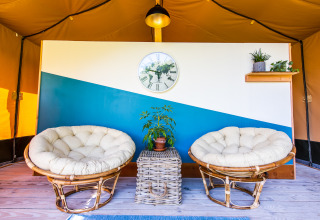 Interno di una tenda glamping con due sedie rotonde, tavolo in vimini, orologio e piante in Auvergne.