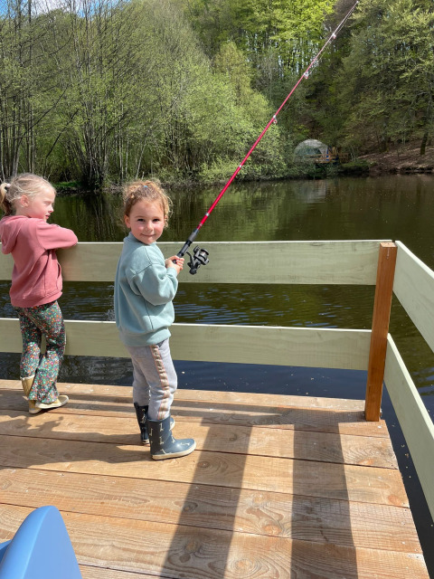 Due bambini pescano su un pontile in legno vicino al lago, con tenda Glamping Auvergne sullo sfondo verde.