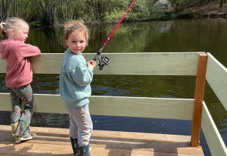 Due bambini pescano su un pontile in legno vicino al lago, con tenda Glamping Auvergne sullo sfondo verde.