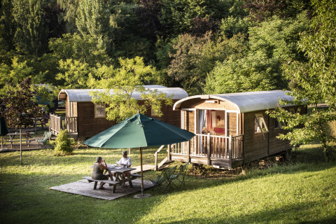 Dos personas se sientan bajo una sombrilla frente a cabañas de glamping en plena naturaleza en Huttopia Sarlat.