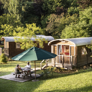 Two people sit under an umbrella at a table near cozy wooden glamping cabins in lush nature at Huttopia Sarlat.