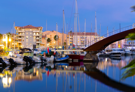 Vista nocturna del puerto deportivo con barcos, alojamiento glamping y edificios iluminados cerca del agua.