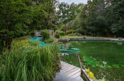 Piscina naturale a Uhlenköper-Camp Uelzen, circondata da vegetazione e uno scivolo d’acqua blu.