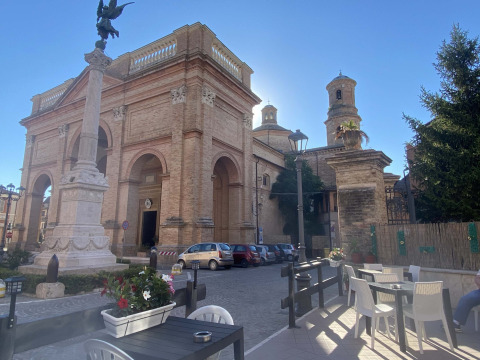 Straataanzicht bij een Italiaanse kerk met buitenterras, bloembakken, geparkeerde auto's en zon.