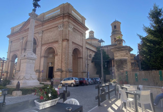 Blick auf eine italienische Kirche mit Freiluftcafé, Sitzplätzen, Blumen und Autos bei Sonne.