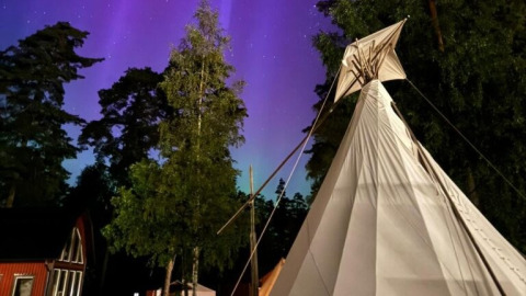 Nordic Woods Wildernis Glamping Zweden: tipitent onder noorderlicht, omgeven door bosrijke natuur in de nacht.